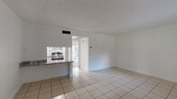 Located at 7655 NW 42nd Pl in Sunrise, Florida, this single-story unit presents a clean, functional layout with a bright, neutral palette throughout. The main living area is generously proportioned, featuring light-toned ceramic tile flooring and a textured ceiling that adds subtle visual interest. White walls and natural light filtering in from a window and nearby balcony access contribute to an open, airy atmosphere. A secondary living or flex space near the entry shares the same tile flooring and benefits from a window overlooking the outdoors.

The kitchen is efficiently designed and opens toward the living area via a peninsula counter finished in granite, providing a practical workspace and casual dining option. White cabinetry with glass-front uppers offers ample storage, while a stainless steel range with overhead ventilation hood anchors the cooking space. Tile flooring continues seamlessly through the kitchen.

The bedroom features light wood-tone laminate flooring and a sliding glass door that leads to a private balcony, allowing for natural light and outdoor access. The bathroom is finished with wood-tone laminate flooring, white shaker-style cabinetry, and a light stone-finish countertop, maintaining the cohesive, clean aesthetic found throughout the space.