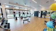 Located at 2200 W Meeker St in Kent, Washington, this single-level fitness center offers a comprehensive and modern workout environment. The space is designed with an open, airy layout featuring light hardwood-style flooring, crisp white walls, and elevated ceilings that contribute to a spacious, energizing atmosphere. Linear LED lighting fixtures and recessed ceiling lights ensure consistent, well-distributed illumination throughout.

Large floor-to-ceiling windows and transom windows line the perimeter walls, flooding the interior with natural light and offering direct views of an outdoor pool and the surrounding community. A full-length mirror framed in yellow accent trim further enhances the sense of openness along one wall.

The cardio zone is well-appointed with multiple treadmills, elliptical trainers, and a rowing machine, all positioned to take advantage of the poolside views. The strength training area includes cable and weight machines, a dedicated dumbbell rack with a range of free weights, and functional training accessories such as stability balls, medicine balls, and resistance bands displayed on organized racks. Decorative athletic silhouette graphics add a dynamic visual element to the strength zone. Glass partitioning subtly defines areas within the space while maintaining an open feel throughout.