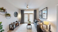 This single-floor residence offers a thoughtfully designed layout that balances functionality with contemporary style. The living room serves as the central gathering space, featuring light wood-style flooring, a ceiling fan, and a glass-paneled door framed by floor-to-ceiling gray drapes that invite natural light while providing privacy. A transom window above the door further brightens the room. Framed artwork and an open shelving unit add character to the space.

The kitchen is well-equipped with white shaker-style cabinetry, granite-look countertops, and black appliances including a range, dishwasher, microwave, and refrigerator. Track lighting illuminates the space, and light wood-style flooring continues throughout, creating visual continuity.

The bedroom is carpeted and features a window with sheer curtains, providing a soft, restful ambiance. Vaulted ceiling details add dimension, and the room accommodates both a sleeping area and a functional desk workspace with floating shelves above.

The bathroom maintains the home's neutral palette with warm beige tones, a shower/tub combination, and tasteful decorative accents. Overall, the residence presents a cohesive, modern aesthetic throughout.