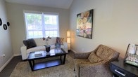 This single-floor space offers a comfortable and thoughtfully designed interior that blends functionality with aesthetic charm. The sitting area serves as the heart of the space, bathed in abundant natural light streaming through a large, white-framed window that frames a lush outdoor view. Neutral gray walls provide a clean, calming backdrop that complements the warm tones found throughout the room. A plush, cream-colored sofa is paired with a patterned accent chair, creating an intimate and inviting seating arrangement centered around a sleek, dark-finished coffee table. A textured area rug anchors the space, adding warmth and depth underfoot. An elegant floor lamp with a decorative column design adds a soft ambient glow, while a vibrant abstract painting mounted on the wall introduces a bold pop of color and artistic personality. Finishing touches such as a potted orchid and tastefully arranged books on the coffee table contribute to the space's refined yet approachable feel. Overall, the space strikes a harmonious balance between comfort and style, making it an ideal environment for relaxation or conversation.