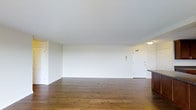This single-floor space presents a straightforward, open layout characterized by clean white walls and warm hardwood flooring that extends throughout the main living area. The floors display rich wood tones with natural grain variation, adding warmth and texture to the otherwise neutral interior palette. The ceiling is smooth and painted white, contributing to a bright, airy atmosphere. The open main area provides generous room to configure furnishings in a variety of arrangements, making it adaptable for different lifestyle needs. A wall-mounted intercom panel is visible, offering a practical communication feature. Toward one side of the space, overhead lighting illuminates what appears to be an adjacent kitchen or entry area, partially visible with a countertop edge in view. Two doorways are present—one on either side of the main wall—providing access to additional areas of the unit. The overall ambiance is clean and minimalist, with neutral tones throughout creating a blank canvas well-suited to a range of interior design styles. The space feels open and uncluttered, emphasizing its functional, move-in-ready character.