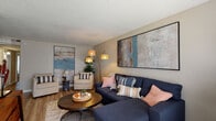 This single-floor residence offers a thoughtfully designed layout that balances comfort and functionality across its key living areas. The living room serves as the centerpiece of the home, featuring warm wood-look flooring, a textured ceiling, and generous wall space showcasing large-format abstract artwork. A deep navy sectional sofa, complementary accent chairs, and a multi-arm arc floor lamp create an inviting and well-lit atmosphere. The adjacent dining area provides dedicated space for shared meals, complete with a wood-top dining table, white chairs, and a bold colorful wall piece adding a vibrant accent. The kitchen is outfitted with crisp white shaker-style cabinetry, light granite-look countertops, a tile backsplash, stainless steel appliances including a dishwasher, and a double-basin sink beneath a window. Both bedrooms feature plush carpeting and ceiling fans. The primary bedroom includes a reach-in closet and direct access to the bathroom, while the secondary bedroom benefits from natural light through a window with blinds. The bathroom is finished with wood-look flooring, white cabinetry with granite-look counters, and a tiled tub-shower combination, presenting a clean and updated aesthetic throughout.