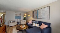 This single-floor residence offers a thoughtfully designed layout that balances comfort and functionality across its key living areas. The living room serves as the centerpiece of the home, featuring warm wood-look flooring, a textured ceiling, and generous wall space showcasing large-format abstract artwork. A deep navy sectional sofa, complementary accent chairs, and a multi-arm arc floor lamp create an inviting and well-lit atmosphere. The adjacent dining area provides dedicated space for shared meals, complete with a wood-top dining table, white chairs, and a bold colorful wall piece adding a vibrant accent. The kitchen is outfitted with crisp white shaker-style cabinetry, light granite-look countertops, a tile backsplash, stainless steel appliances including a dishwasher, and a double-basin sink beneath a window. Both bedrooms feature plush carpeting and ceiling fans. The primary bedroom includes a reach-in closet and direct access to the bathroom, while the secondary bedroom benefits from natural light through a window with blinds. The bathroom is finished with wood-look flooring, white cabinetry with granite-look counters, and a tiled tub-shower combination, presenting a clean and updated aesthetic throughout.