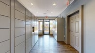 This single-level package and mail room offers a well-organized, corridor-style layout designed to efficiently manage parcel deliveries. The space is lined on both sides with rows of smart parcel lockers finished in a neutral gray, stacked floor to ceiling and extending the full length of the room, providing substantial package storage capacity. A traditional mailbox unit is also present along one wall, accommodating standard mail distribution alongside the automated locker system.

The flooring throughout consists of wood-look planks in a warm, medium-toned finish, lending a polished yet durable aesthetic appropriate for a high-traffic utility space. Recessed ceiling lights provide consistent, even illumination across the corridor. The walls are painted in a soft blue-gray tone that complements the cool metallic finishes of the locker units.

At one end of the room, a glass-panel exterior door with dark metal framing allows natural light to filter into the space while offering a view of the surrounding outdoor courtyard, which features brick paving and landscaping. Additional windows enhance the brightness of the entry area. A secondary interior door provides access to adjacent areas of the building. The overall design is clean, functional, and purposefully laid out for convenient resident package retrieval.