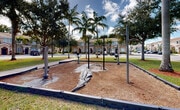 Located at 1140 SE 24th Rd in Homestead, Florida, this outdoor community recreation space offers a well-appointed fitness and play area set within a beautifully landscaped residential neighborhood. The space is defined by a wood chip-surfaced fitness zone bordered by low edging, housing multiple open-air exercise stations designed for strength and flexibility training. Tall, mature royal palms and broad shade trees are strategically positioned throughout the area, providing natural canopy and a distinctly tropical Florida ambiance. Lush green lawns surround the activity zones, complemented by red mulch landscaping beds that add visual contrast and texture. A children's playground with colorful equipment, including a slide, is visible nearby, making this a versatile recreational destination for all ages. Wide concrete pathways connect the various areas, ensuring easy accessibility throughout the space. Street lamps are positioned across the grounds to support use during evening hours. The backdrop features a row of Mediterranean-style two-story residential buildings with terracotta tile roofs, arched entryways, and warm stucco facades, reinforcing the cohesive character of the community. The open sky and abundant natural light enhance the inviting, active atmosphere of this outdoor amenity space.