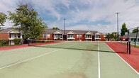 This outdoor tennis court is a well-defined recreational amenity situated within a multi-building residential community. The court features a classic green hard-surface playing area with crisp white boundary lines, contrasted by a red surround that clearly delineates the out-of-bounds zones. A regulation net is installed at the center, and tall floodlight poles are positioned to enable play during evening hours. The entire court is enclosed by a chain-link perimeter fence, ensuring both safety and ball containment during play.

The surrounding environment enhances the outdoor experience, with mature trees providing natural shade and greenery along the court's edges. Views from the court reveal the broader community grounds, including a pool area with lounge seating, a covered gazebo, and brick residential buildings with balconies and pitched rooflines. The landscaped grounds feature open lawn areas that contribute to the open, park-like atmosphere.

While the court surface shows visible weathering and crack lines consistent with age, the overall layout and infrastructure remain functional and clearly defined. The combination of the court, pool, and gazebo points to a community amenity area designed for active outdoor recreation and leisure, set against a backdrop of established residential architecture and lush tree canopy.