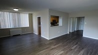 This single-floor residence offers a thoughtfully designed open-concept layout that maximizes both space and functionality. The expansive main living area is finished with rich, dark hardwood flooring that extends seamlessly throughout, lending a warm and cohesive feel to the interior.

A defining architectural feature of the space is the pass-through window cut into the central dividing wall, which connects the kitchen to the main living area. This detail enhances the open feel while maintaining a subtle separation between the cooking and living zones. The kitchen is equipped with warm-toned wood cabinetry and black appliances, including a built-in microwave, providing a classic and functional cooking environment.

The neutral gray walls throughout the space offer a clean, modern backdrop that complements the warm flooring tones. Large vertical blinds on the windows allow for flexible light control, filling the space with natural light when open. Additional doorways visible from the main living area suggest access to further rooms, contributing to the overall sense of a well-connected and practical floor plan. Clean white baseboards and trim complete the interior, adding a crisp finishing touch to the overall aesthetic.