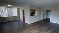 This single-floor residence offers a thoughtfully designed open-concept layout that maximizes both space and functionality. The expansive main living area is finished with rich, dark hardwood flooring that extends seamlessly throughout, lending a warm and cohesive feel to the interior.

A defining architectural feature of the space is the pass-through window cut into the central dividing wall, which connects the kitchen to the main living area. This detail enhances the open feel while maintaining a subtle separation between the cooking and living zones. The kitchen is equipped with warm-toned wood cabinetry and black appliances, including a built-in microwave, providing a classic and functional cooking environment.

The neutral gray walls throughout the space offer a clean, modern backdrop that complements the warm flooring tones. Large vertical blinds on the windows allow for flexible light control, filling the space with natural light when open. Additional doorways visible from the main living area suggest access to further rooms, contributing to the overall sense of a well-connected and practical floor plan. Clean white baseboards and trim complete the interior, adding a crisp finishing touch to the overall aesthetic.