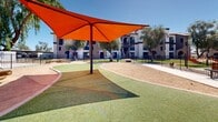 Located at 4722 E Bell Rd in Phoenix, Arizona, this thoughtfully designed outdoor amenity space serves as a dedicated dog park and recreational area set within a residential community. The grounds are enclosed by black iron fencing, providing a secure environment for pets to roam freely. The space is divided into distinct zones, including a sandy off-leash run area equipped with agility features such as a log balance beam and other interactive elements, offering dogs both physical and mental stimulation. A vibrant synthetic turf area winds through the park, offering a clean and comfortable surface for play, and is complemented by a striking orange shade canopy that provides welcome relief from the Arizona sun. String lights are strung overhead in one section of the grounds, adding a welcoming ambiance during evening hours. A covered pergola structure serves as a central gathering point, surrounded by mature trees including palms and olive trees that lend natural shade and visual character to the space. The surrounding architecture reflects a Spanish-Mediterranean style, with white stucco buildings, terracotta roof tiles, and arched detailing that frame the outdoor area beautifully. The open sky and abundant natural light enhance the overall appeal, making this a vibrant and functional outdoor destination for residents and their pets.