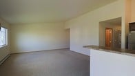 This single-floor residence offers a clean, functional layout designed to maximize usability and comfort. The main living area is finished with wall-to-wall carpet in a neutral tone, providing a soft and inviting foundation that complements the bright, white walls throughout. The open-concept design creates a generous sense of space, allowing natural light to permeate the room and contribute to an airy, welcoming ambiance. A defining architectural feature is the built-in breakfast bar, topped with a granite-style surface and set into a crisp white half-wall that separates the living area from the adjoining kitchen. This pass-through design encourages connectivity between spaces while maintaining a sense of distinction between the living and kitchen areas. The kitchen itself is accessible through an open archway, offering a seamless flow between the two spaces. The neutral color palette of whites and warm beiges throughout creates a versatile backdrop suitable for a variety of interior styles. The overall layout is straightforward and practical, with smooth walls, consistent flooring, and a cohesive design language that makes the space feel unified and easy to personalize.