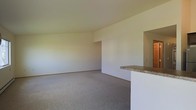 This single-floor residence offers a clean, functional layout designed to maximize usability and comfort. The main living area is finished with wall-to-wall carpet in a neutral tone, providing a soft and inviting foundation that complements the bright, white walls throughout. The open-concept design creates a generous sense of space, allowing natural light to permeate the room and contribute to an airy, welcoming ambiance. A defining architectural feature is the built-in breakfast bar, topped with a granite-style surface and set into a crisp white half-wall that separates the living area from the adjoining kitchen. This pass-through design encourages connectivity between spaces while maintaining a sense of distinction between the living and kitchen areas. The kitchen itself is accessible through an open archway, offering a seamless flow between the two spaces. The neutral color palette of whites and warm beiges throughout creates a versatile backdrop suitable for a variety of interior styles. The overall layout is straightforward and practical, with smooth walls, consistent flooring, and a cohesive design language that makes the space feel unified and easy to personalize.