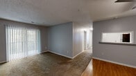 This single-floor residence is thoughtfully laid out, offering a comfortable and functional living environment. The main living area is generously proportioned, featuring warm neutral-toned carpet underfoot and large vertical blind-covered sliding doors that allow natural light to filter through. A wall-mounted pass-through window connects the living space to an adjacent area, adding an open, communicative feel between rooms.

The flooring transitions from carpet in the living and bedroom areas to hardwood-style planks throughout the kitchen and hallway. The kitchen is equipped with light wood cabinetry, light-toned countertops, and a full-size stainless refrigerator, providing practical storage and workspace.

A central hallway connects multiple rooms, with several doors visible in succession, suggesting well-defined private spaces. The bedrooms are carpeted, neutral in tone, and receive natural light through windows fitted with horizontal blinds. The primary bedroom benefits from direct access to an en-suite bathroom, which features a full-size soaking tub with a tub-shower combination and chrome fixtures.

Throughout, textured ceilings, white baseboards, and warm gray walls create a consistent and clean aesthetic. Baseboard heating units are present in several rooms.