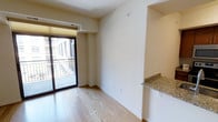 This single-floor residence offers a clean, modern aesthetic defined by light hardwood flooring, neutral wall tones, and an efficient open layout. The main living area is filled with natural light, thanks to a large sliding glass door that opens onto a private balcony enclosed by a classic metal railing — providing a pleasant outdoor extension of the interior space and views of the surrounding building exterior. A built-in granite countertop peninsula serves as a practical transition between the living and kitchen areas, offering additional prep or dining surface space while contributing a polished, upscale detail to the overall design. Recessed HVAC ventilation is thoughtfully integrated near the ceiling, ensuring comfort without disrupting the streamlined look of the space. Neutral cream-toned walls throughout create a versatile backdrop well-suited for a variety of interior styles. The layout balances openness with defined functional zones, making efficient use of the available footprint. Overall, the space presents a well-appointed, move-in-ready environment with quality finishes and practical design elements that cater to everyday modern living.