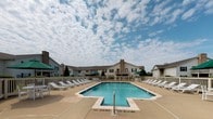 Located at Fairfield Place in O'Fallon, Illinois, this community pool facility offers a well-maintained outdoor recreational area designed for relaxation and leisure. The centerpiece is a generously sized swimming pool with crystal-clear blue water, clearly marked with no-diving indicators along its edges, and surrounded by a wide concrete deck that accommodates numerous white lounge chairs and green-accented patio umbrellas for shade. The pool area is enclosed by a white picket-style fence, providing both safety and a clean aesthetic.

Access to the pool is controlled through a gated entry, prominently displaying posted pool rules and regulations to ensure the safety of all users. A covered walkway runs along the perimeter of the facility, supported by decorative columns with brick bases and featuring a white-paneled ceiling. This shaded corridor provides a comfortable transition between the interior amenity building and the pool deck. The amenity building itself features teal-accented entry doors with sidelight windows, white siding with dark shutters, and well-kept landscaping with neatly trimmed shrubs. Security is supported by surveillance cameras positioned at the entrance. The surrounding residential buildings are visible beyond the fence, reinforcing the community-centered nature of this outdoor amenity.
