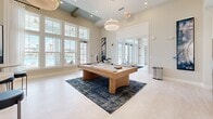 Located in Santa Rosa Beach, Florida, this sophisticated single-floor clubhouse offers a thoughtfully designed living and gathering space that blends coastal elegance with refined comfort. The interior is anchored by soaring ceilings adorned with exposed wood beam accents, lending a warm, architectural character to the open layout. Large floor-to-ceiling windows and French doors flood the space with natural light while framing views of the surrounding pool area and lush Florida landscape.

The main lounge area features a generously sized sectional sofa and woven accent chairs arranged around a sculptural geometric coffee table, all set atop a bold navy chevron-patterned area rug. A wall-mounted flat-screen television serves as a functional focal point on one side of the room. Complementing the lounge, a dedicated billiards area showcases a sleek hardwood pool table, providing an inviting recreational centerpiece.

A standout design feature is the pair of built-in banquette dining nooks, each framed by dramatic floor-to-ceiling marble-look accent walls in soft grey and blue tones, illuminated by woven pendant lights. The light wood flooring, neutral color palette, and curated coastal furnishings throughout create a cohesive, airy ambiance that reflects the relaxed yet upscale character of the Santa Rosa Beach lifestyle.