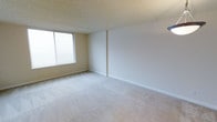 This single-floor space presents a clean, minimalist interior with a straightforward layout. The main living area is finished with wall-to-wall neutral beige carpeting, providing a soft underfoot feel that extends throughout the room. Walls are painted in a light, warm off-white tone, contributing to an open and airy atmosphere.

A large window along one wall allows generous natural light to fill the room, fitted with horizontal blinds for adjustable light control and privacy. The textured popcorn ceiling adds a subtle dimensional quality overhead, a classic finishing detail found in many buildings of this style.

White baseboards run along the perimeter of the room, neatly framing the carpet and walls. The room's proportions offer a comfortable sense of open space, with enough room to accommodate a variety of furniture arrangements. The overall ambiance is calm and neutral, providing a blank canvas well-suited for personalization. The space is practical and functional, reflecting a straightforward residential design with attention to basic comfort and livability.