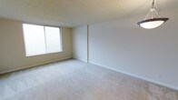 This single-floor space presents a clean, minimalist interior with a straightforward layout. The main living area is finished with wall-to-wall neutral beige carpeting, providing a soft underfoot feel that extends throughout the room. Walls are painted in a light, warm off-white tone, contributing to an open and airy atmosphere.

A large window along one wall allows generous natural light to fill the room, fitted with horizontal blinds for adjustable light control and privacy. The textured popcorn ceiling adds a subtle dimensional quality overhead, a classic finishing detail found in many buildings of this style.

White baseboards run along the perimeter of the room, neatly framing the carpet and walls. The room's proportions offer a comfortable sense of open space, with enough room to accommodate a variety of furniture arrangements. The overall ambiance is calm and neutral, providing a blank canvas well-suited for personalization. The space is practical and functional, reflecting a straightforward residential design with attention to basic comfort and livability.
