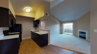 This single-story residence is defined by its striking vaulted ceilings, which lend a sense of openness and volume to the main living areas. The open-concept layout connects the kitchen and living room seamlessly, creating a functional and cohesive space. The kitchen is outfitted with dark espresso cabinetry, light countertops, black appliances including a refrigerator, range, and microwave, and a stainless steel sink. A peninsula with a breakfast bar provides a natural divide between the kitchen and the adjacent dining area, which features hardwood-style flooring and a pendant light fixture overhead.

The living room is carpeted and anchored by a fireplace, adding warmth to the space. A sliding glass door leads to a private balcony, allowing natural light to flow in and offering outdoor views. The bedroom is generously sized with plush carpeting, a closet, and direct access to the hallway. The bathroom includes dark cabinetry with a light countertop vanity, a framed glass walk-in shower, hardwood-style flooring, and a grab bar. Throughout, neutral wall tones and consistent finishes create a cohesive and practical living environment.