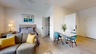 Located at 180 N Martin Luther King Blvd in Lexington, Kentucky, this single-floor studio unit offers a thoughtfully designed open-plan layout that efficiently combines living, sleeping, dining, and cooking areas into one cohesive space. Light wood-style flooring flows throughout, complementing the clean white walls and creating a bright, contemporary ambiance.

The living area features a ceiling fan with light fixture, a wall-mounted flat-screen TV, and a large window fitted with blinds that allows natural light to fill the room. A comfortable sofa, coffee table, and dresser complete the living zone. Adjacent to it, the sleeping area is seamlessly integrated within the open floor plan.

The dining area includes a compact table with teal modern chairs, providing a defined space for meals. The kitchenette is equipped with warm wood cabinetry, stainless steel appliances including a refrigerator, electric range, and microwave, along with open shelving and a single-basin sink with ample counter space.

The private bathroom is well-appointed with a wood-tone vanity, white countertop, a framed mirror with overhead lighting, and a shower-tub combination. A walk-in closet provides convenient storage. The unit's efficient design and modern finishes make it a functional and comfortable urban living space in downtown Lexington.