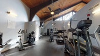 This fitness center combines striking architectural character with a thorough range of exercise equipment. The space is defined by its dramatic vaulted ceiling clad in warm wood paneling, supported by exposed timber beams and arched accents that lend a distinctive, welcoming atmosphere. Ceiling fans are suspended throughout, ensuring comfortable airflow during workouts. High clerestory windows line the upper walls, drawing in generous natural light and contributing to an open, airy environment.

The rubber-floored workout area is organized to accommodate both cardio and strength training. Cardio equipment includes multiple treadmills, elliptical trainers, stationary bikes, and stair climbers positioned along the perimeter and central zones. The strength training section features a Life Fitness cable machine, a multi-station functional trainer, a dumbbell rack with a full set of free weights, and adjustable benches. Large wall-mounted mirrors extend across one side, visually expanding the space and allowing users to monitor their form. Wall-mounted televisions provide entertainment during workouts. A dedicated area with a green accent wall offers additional open floor space, suitable for stretching or functional training, complemented by mounted wall storage for accessories such as medicine balls.