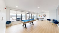 This contemporary recreation room is thoughtfully designed for entertainment and leisure, combining a clean modern aesthetic with a versatile layout. The space is anchored by light-toned hardwood-style flooring that runs throughout, complementing the crisp white walls and recessed square lighting overhead that ensures the room is evenly and brightly lit.

At the center of the main area sits a striking pool table with a vivid blue felt surface and a natural wood and black metal frame, serving as the focal point of the room. Nearby, a ping-pong table offers additional recreational options, making the space well-suited for social gatherings. A dedicated lounge zone features a row of plush navy-blue velvet chairs with gold-toned legs arranged against a teal accent wall, below a wall-mounted flat-screen television — creating an inviting area to relax and watch.

Large floor-to-ceiling windows and glass doors line one side of the room, flooding the interior with natural light and offering sweeping views of the surrounding landscape and hills. These doors open directly onto an outdoor terrace, which is equipped with seating and a hot tub, seamlessly extending the living and entertainment experience outdoors. A decorative mirror and colorful wall art add personality to the space, while a wall-mounted mini-split unit ensures year-round comfort.