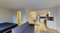 This single-floor space showcases a thoughtfully designed open-concept layout that seamlessly connects the living area and kitchen. The living area features a comfortable sectional sofa and light hardwood-style flooring that extends throughout the space, creating a cohesive and airy feel. Recessed lighting illuminates the interior, complementing the neutral light-blue and white wall tones.

The kitchen is well-appointed with white shaker-style cabinetry, a dark slate tile backsplash, stainless steel appliances including a refrigerator and range, and a sink positioned along the perimeter wall. A small kitchen island with bar seating provides additional functionality and separates the kitchen from the living space.

A hallway visible from the main living area leads to additional rooms, with doors suggesting at least one bedroom and a closet space, offering a clear sense of a functional multi-room layout. The consistent flooring and clean architectural lines give the entire space a modern, unified aesthetic. The recessed lighting and bright white ceilings enhance the overall sense of openness throughout this efficiently designed interior.