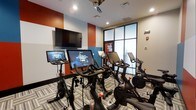 The Spin Room is a purpose-built indoor cycling studio designed to deliver an immersive fitness experience. The space features a fleet of Peloton stationary bikes, each equipped with its own interactive touchscreen display, allowing riders to access live and on-demand classes at any time. A wall-mounted flat-screen television at the front of the room provides an additional viewing option, supporting group or instructor-led sessions.

The room's interior design is energetic and motivating, with bold accent walls in deep orange and teal contrasting against a neutral gray base. Recessed lighting illuminates the space evenly from above, while a large glass partition wall on one side introduces natural light and a sense of visual openness without compromising the focused studio atmosphere.

Patterned, striped carpeting covers the floor throughout, adding texture and a degree of sound absorption suited to a high-energy workout environment. The layout accommodates multiple bikes arranged to provide each user with adequate space and clear sightlines to the screens. Overall, the Spin Room combines modern fitness technology with a dynamic, studio-style ambiance.