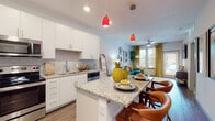 This single-story space showcases a thoughtfully designed open-concept layout that seamlessly connects the kitchen, dining, and living areas. The kitchen is a standout feature, outfitted with crisp white cabinetry, granite countertops with subtle veining, a classic subway tile backsplash, and a full suite of stainless steel appliances. A central island with bar seating serves as both a functional workspace and a social hub, accented by bold red pendant lights that add a striking visual element. Wood-look flooring runs throughout the main living areas, lending warmth and continuity to the space.

The living room is bright and inviting, benefiting from natural light through large windows dressed with layered curtains and blinds. A ceiling fan provides comfort overhead, while the room's layout accommodates comfortable seating and décor.

The bedrooms are carpeted for comfort and each receive natural light from well-positioned windows. One bedroom includes access to a spacious walk-in closet fitted with wire shelving, providing generous storage capacity. Throughout the space, the design balances modern, clean-lined finishes with warm, eclectic styling that gives the unit a welcoming and distinctive ambiance.