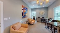 This well-appointed single-floor space combines comfort and functionality through a series of thoughtfully designed rooms. The main lounge area serves as an inviting focal point, featuring plush seating including patterned armchairs and a sofa in warm orange tones, set against cool grey walls. A bold, large-scale abstract painting in vibrant blues, oranges, and reds commands attention on the wall, adding a strong artistic statement to the room. A sleek modern chandelier overhead provides ambient lighting, while crown molding along the ceiling lends an elegant, finished touch.

An adjacent business center offers a productive workspace with a built-in desk spanning the width of the room, outfitted with multiple workstations, a printer, and ample drawer storage beneath. Soft blue-grey walls and carpet flooring create a calm, focused atmosphere, complemented by a decorative sunburst mirror and natural light filtering through horizontal blinds. A bright orange accent chair adds a pop of color to the otherwise serene workspace.

A separate dining nook provides an intimate setting with a round glass-top table and upholstered chairs featuring oval back cutouts. A window overlooking lush greenery fills the space with natural light. Together, these areas form a cohesive, stylish environment ideal for both relaxation and productivity.