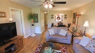 This single-floor home presents an inviting open-concept layout that seamlessly connects the living and dining areas. The living room is anchored by warm hardwood flooring that extends throughout the space, complemented by neutral beige walls that create a welcoming atmosphere. A ceiling fan with a light fixture overhead provides both function and a casual charm to the room.

The living area is thoughtfully arranged, featuring a plush loveseat accented with decorative pillows and a patterned area rug in rich red tones. A coffee table and accompanying furniture complete the seating arrangement. A decorative pedestal with a lush potted plant adds a touch of greenery and elegance to the corner near the entryway.

Just beyond the living area, the dining space is distinguished by an elegant chandelier that casts warm light over a dark wood dining set. Wall-mounted decorative accents and sconces frame the dining area, adding architectural interest. A doorway visible from the living room suggests additional rooms beyond, providing a sense of depth and flow throughout the home. The overall ambiance is warm, classic, and well-appointed.