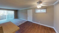 This single-floor residence offers a functional and comfortable layout with a cohesive neutral color palette of warm taupe walls, white trim, and textured ceilings throughout. The open living and dining area is finished with hardwood-style flooring, crown molding, a ceiling fan with integrated lighting, and a large window fitted with vertical blinds that allows generous natural light to filter in. A sliding glass door provides access to an outdoor area, extending the sense of space. The kitchen features white cabinetry, a countertop peninsula, a dishwasher, refrigerator, and a sink beneath under-cabinet lighting, making it both practical and well-appointed. The bedrooms are carpeted and bright, each equipped with large windows dressed in vertical blinds that offer views of mature trees and greenery outside. The space includes two bathrooms: one with a tub-shower combination, white vanity cabinetry, and hardwood-style flooring, and a second with a glass-door walk-in shower, a vanity with a wide mirror, and matching finishes. The overall layout, as visible in the floorplan, is efficiently organized to maximize usability across all living areas.