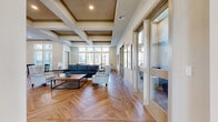 This thoughtfully designed community space spans a generous single-floor footprint, offering a versatile and welcoming environment for residents. The open-plan layout flows seamlessly between a comfortable lounge area, a dining area, and a dedicated activity room, all connected by striking herringbone-patterned hardwood flooring that runs throughout.

The lounge area is anchored by a coffered ceiling with warm-toned beams that add architectural depth and character. Plush seating, including a navy blue sofa and tufted armchairs, is arranged around a central coffee table, creating an inviting gathering space. Framed botanical artwork adorns the neutral-toned walls, contributing to the space's refined, welcoming ambiance.

Floor-to-ceiling windows and transom windows wrap around much of the space, flooding the interior with abundant natural light and offering views of the surrounding landscape. The dining area features multiple round and rectangular tables with cross-back chairs, illuminated by sleek pendant lights and backed by a decorative tile accent wall with a stainless steel refrigerator and cabinetry.

The activity room, accessible via glass-paneled sliding doors, provides a flexible, light-filled space for organized events. The overall design balances modern elegance with functional comfort, making it an ideal communal hub.