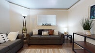 This single-floor unit offers a thoughtfully designed layout with multiple bedrooms, two full bathrooms, a living area, and a functional kitchen. The living room serves as the central gathering space, featuring warm hardwood-style flooring, crown molding, and a large window with blinds that allows natural light to fill the room. A full-size sofa and loveseat provide ample seating, complemented by a cozy area rug and accent lighting. The entryway flows naturally into the living space, featuring a wall sconce and a decorative mirror that add a welcoming touch. The galley-style kitchen is equipped with gray cabinetry, light countertops, and a full suite of white appliances including a stove, microwave, and refrigerator. Hardwood-style flooring and a window over the counter keep the kitchen bright and practical. Each bedroom features plush carpeting, a dedicated study desk, and an open closet with hanging space — ideal for student living. The bathrooms are finished with wood-look flooring, gray vanity cabinetry, light countertops, and a shower-tub combination, offering a clean and functional design throughout. The overall ambiance is casual, comfortable, and well-suited for everyday living.
