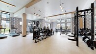 This contemporary fitness center is thoughtfully designed to accommodate a wide variety of training needs, both indoors and outdoors. The interior gym is spacious and airy, featuring soaring ceilings with warm wood beam accents and sleek linear pendant lighting that illuminates the space efficiently. The floors are finished in a durable speckled rubberized material, ideal for high-impact activity. Bold geometric-patterned wallcoverings add a stylish visual contrast against the crisp white ceilings and structural columns clad in natural wood tones. Large black-framed windows line the walls, flooding the interior with natural light and offering views of the surrounding outdoor grounds and pool area. Large mirrors further enhance the sense of openness throughout. The fitness floor is comprehensively outfitted with strength training machines, a multi-station cable system, a full dumbbell rack, adjustable benches, treadmills, ellipticals, stationary bikes, and a rowing machine. A flat-screen television is mounted for entertainment. Glass doors open directly to an outdoor training zone, which features a fully loaded functional fitness rig equipped with battle ropes, kettlebells, medicine balls, weight plates, and suspension trainers — all set against an open-air backdrop with views of the surrounding community.