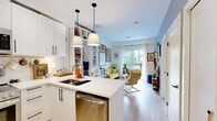 This well-designed one-bedroom, one-bathroom unit on a single floor offers a smart and efficient layout that maximizes every square foot. The open-concept kitchen and living area serves as the heart of the home, featuring white shaker-style cabinetry with matte black hardware, stainless steel appliances including a full-size refrigerator, gas range, and over-the-range microwave, as well as a kitchen island with a clean white countertop. Pendant lighting and track lighting illuminate the space, complementing the light-toned hardwood-style flooring throughout. The living area is bright and airy, highlighted by large sliding glass doors that open onto a private balcony, filling the room with natural light. Built-in white shelving along one wall provides ample storage and display space. The bedroom is a comfortable retreat, featuring a built-in wall unit with integrated shelving and cabinetry, a window with exterior views, and soft carpet underfoot. The bathroom offers a full tub-shower combination, a vanity with storage, and light tile flooring. Throughout the unit, a cohesive white palette with warm wood accents creates a clean, modern, and inviting atmosphere.