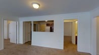 This single-floor residence is designed with a practical and functional open layout that promotes easy flow between living spaces. The main living area features wall-to-wall carpet and is finished with classic crown molding that runs along the ceiling, adding a clean architectural detail throughout. A flush-mount ceiling light provides overhead illumination in the living space.

The kitchen is positioned adjacent to the living area and is accessible via a pass-through opening in the half-wall partition, creating a sense of connectivity while maintaining a defined boundary between the two spaces. The kitchen is equipped with dark wood cabinetry, a white refrigerator, and appears to include additional pantry or closet storage. The pass-through counter provides a convenient serving or interaction point between the kitchen and living area.

From the living room, doorways lead to additional rooms. One door on the left opens to a closet or utility space, while an opening on the right leads to what appears to be a bedroom or additional living area, accompanied by a well-lit bathroom. The neutral color palette of white walls, beige carpet, and warm wood tones throughout creates a cohesive, understated aesthetic that is both welcoming and easy to personalize.