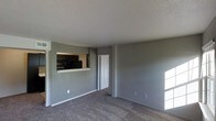 This single-floor space presents a straightforward and functional layout designed for comfortable everyday living. The living area is finished with wall-to-wall carpet in a neutral tone, complementing the cool grey walls that lend the space a clean, understated aesthetic. A textured ceiling adds subtle depth overhead, while ample natural light filters in, brightening the interior.

A standout architectural feature is the pass-through window that connects the living area to the kitchen, creating a sense of openness between the two spaces. Through the pass-through, black appliances and dark countertops are visible, offering a contrast to the lighter tones of the living area. This design element enhances the flow and connectivity of the layout without fully opening the two rooms to one another.

A white paneled door provides access to an adjacent room or hallway, contributing to the clean, minimal character of the interior. Electrical outlets are conveniently placed along the baseboards throughout the space. The overall ambiance is calm and neutral, providing a versatile backdrop that can be easily personalized. The layout is efficient and well-organized, making practical use of the available space across the single floor.