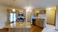 This single-floor space centers around a thoughtfully designed kitchen that serves as its standout feature. The kitchen is outfitted with a full suite of stainless steel appliances, including a refrigerator, electric range with microwave above, and a dishwasher, providing a modern and functional cooking environment. Warm-toned wood cabinetry lines the walls, offering generous storage throughout, while a dedicated walk-in corner pantry adds exceptional organizational capacity. A granite-topped kitchen island anchors the space, providing additional prep area and a natural gathering point. Pendant lighting hangs above the island, complementing the overall warm ambiance of the room. The sink area is neatly positioned along a secondary counter run, complete with matching cabinetry below and upper cabinets above. Durable hardwood-style flooring extends across the kitchen and adjoining areas, tying the space together with a cohesive, welcoming aesthetic. The open layout between the kitchen and the adjacent living area promotes a comfortable flow, making the space well-suited for both everyday living and entertaining. The neutral wall tones and consistent finishes throughout contribute to a clean, inviting atmosphere.