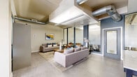 Located at 770 N Logan St in Denver, CO, this clubroom and BBQ patio serves as a well-appointed communal amenity space designed with a contemporary urban aesthetic. The interior features an open lounge area with polished concrete flooring, exposed industrial-style ductwork along the ceiling, and abundant natural light streaming through floor-to-ceiling glass walls and doors. Comfortable seating arrangements, including plush sofas and leather accent chairs, are centered around a round wooden coffee table, creating an inviting gathering environment. A glass-enclosed meeting or conference room with frosted privacy panels offers a more focused workspace adjacent to the main lounge. A wall-mounted television and a rustic wood console table add functional and decorative elements throughout the space. A standout feature is the perforated metal staircase with a marble-topped bar ledge, adding an industrial-chic character to the layout. Package lockers are conveniently integrated near the patio entrance. The outdoor BBQ patio, accented by bold red ceilings and sleek metal railings, offers open-air seating with bar-height chairs, a gas grill, and sweeping views of the surrounding Denver neighborhood, making it an ideal space for social gatherings and relaxation.