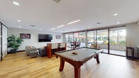 This single-floor community recreation and lounge space is thoughtfully designed for both entertainment and relaxation. The open layout features warm hardwood-style flooring throughout, clean white walls, and recessed lighting complemented by linear LED fixtures that create a bright, welcoming atmosphere.

At the heart of the space sits a full-size billiard table with rich wood detailing, making it a natural focal point for social gatherings. Adjacent to the game area, a built-in cabinetry unit with dark wood finishes houses a popcorn machine and additional storage, adding a fun, amenity-rich touch to the room.

The lounge area is furnished with comfortable gray sofas and accent chairs, accented with colorful throw pillows, and centered around a low-profile coffee table. A large wall-mounted flat-screen TV serves this seating zone, ideal for group viewing. Decorative indoor palms add a lush, organic element to the space.

Large sliding glass doors line one wall, flooding the interior with natural light and offering sweeping views of a beautifully maintained outdoor courtyard. The patio features stone pavers, raised garden beds with seasonal plantings, umbrella-shaded dining tables, and open seating areas — seamlessly extending the living space outdoors. A dining table positioned near the glass doors creates an inviting transition between the indoor and outdoor environments.