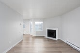 This single-floor residence offers a clean, functional layout designed for comfortable everyday living. The living room serves as the heart of the space, featuring a tile-surround fireplace with a classic black insert, light wood-look flooring, and a baseboard heating system. Large windows with shuttered blinds allow natural light to fill the room, with views of the surrounding exterior. The galley-style kitchen is efficiently designed with ample white cabinetry running floor to ceiling on both sides, complemented by a neutral quartz-style countertop, a tile backsplash, and a stainless steel sink. Black appliances, including an electric range and refrigerator, provide a striking contrast to the bright cabinetry, while track lighting overhead ensures excellent visibility. The bedroom features soft carpet underfoot, a large window with natural tree views, and a baseboard heater. An additional room with light wood flooring includes a built-in closet, providing flexible use as a second bedroom or den. The bathroom is well-appointed with a full soaking tub and shower combination, white tile surround, updated fixtures, a toilet, and a vanity with a light countertop. Throughout the space, a consistent neutral color palette and light flooring create a cohesive and inviting ambiance.