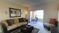 This single-floor space offers a thoughtfully designed living area that balances comfort and style. The living room serves as the heart of the space, featuring plush seating with neutral-toned upholstery accented by rich, dark throw pillows. A decorative framed mirror mounted on the warm beige wall adds depth and elegance, while an ornate wall sconce provides a refined finishing touch. A floor lamp provides soft, ambient lighting to complement the room's inviting atmosphere. The living area is anchored by a geometric-patterned area rug and a sleek dark coffee table, creating a cohesive and well-curated aesthetic. Large sliding glass doors flood the space with abundant natural light and open directly onto a private outdoor patio, seamlessly connecting the interior to the exterior. The patio is furnished with a bistro-style table and ornate chairs set against a backdrop of lush greenery, making it an ideal spot for relaxing or dining al fresco. The carpeted flooring throughout adds warmth and comfort underfoot. Overall, the space presents a harmonious blend of functional design and welcoming ambiance, making it both livable and visually appealing.