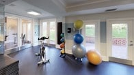 This fitness center is thoughtfully designed to accommodate a wide range of workout needs within a single-floor layout. The space features a two-tone blue and white color scheme on the walls, complemented by dark rubber flooring that provides durability and traction throughout. Recessed panel lighting and ceiling fans ensure the room remains well-lit and comfortable during use.

The cardio section is lined with multiple elliptical trainers and stair climbers, including StairMaster and Magnum branded machines, positioned along a low dividing partition. Rows of windows along the perimeter flood the space with natural light and offer views of the surrounding greenery, creating an energizing atmosphere.

The strength training area houses a Hoist leg press, a multi-station cable machine, a squat rack with weight plates, and additional selectorized equipment. A wall of mirrors enhances the sense of space and supports proper form during exercise.

A dedicated functional training zone near the glass French doors includes a spin bike, medicine ball rack, and a collection of stability balls in various sizes. The doors open to an outdoor patio, providing easy access to fresh air and further extending the welcoming, active ambiance of the facility.