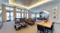 This expansive clubroom is designed for both socializing and leisure, offering a variety of distinct activity zones within a single, open-concept layout. The main lounge area serves as the focal point, anchored by a striking stone-clad fireplace wall with a linear gas fireplace and a mounted flat-screen television. Plush sectional sofas in warm earth tones, accented with mustard yellow throw pillows, are arranged around a tufted coffee table, creating a comfortable gathering space. A wood-paneled tray ceiling with ambient cove lighting adds warmth and architectural interest overhead.

The entertainment amenities are generous and diverse, including a pool table, shuffleboard table, and a dedicated poker table area, all bathed in natural light from oversized floor-to-ceiling windows and sliding glass doors that open to an outdoor terrace with pool views. Sheer draped curtains frame the expansive glazing throughout.

A well-appointed catering kitchen features granite countertops, warm wood cabinetry, subway tile backsplash, and stainless steel appliances. A separate dining area with a large communal table and contemporary pendant lighting completes the space, making it well-suited for private events and community gatherings alike.