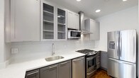This thoughtfully designed kitchen showcases a clean, contemporary aesthetic through a carefully curated combination of materials and finishes. The two-tone cabinetry arrangement pairs light gray upper cabinets — some featuring glass-front panels for open display storage — with darker charcoal lower cabinets, creating a striking visual contrast. Sleek bar-pull hardware ties the look together with a modern sensibility.

The crisp white quartz countertops provide ample prep space and are complemented by a classic white subway tile backsplash that runs the full length of the walls, lending a bright, airy quality to the space. Recessed ceiling lighting, along with pendant light fixtures, ensures the kitchen is well-lit throughout.

The full suite of stainless steel appliances includes a gas range with a built-in oven, a stainless steel microwave tucked neatly beneath the upper cabinets, a matching dishwasher, and a full-size refrigerator. A stainless steel single-basin undermount sink with a contemporary high-arc faucet anchors the workspace. A wall-mounted stainless steel range hood serves as both a functional and stylish focal point above the stove. Rich hardwood flooring grounds the space and adds warmth to the otherwise cool-toned palette.