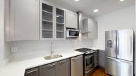 This thoughtfully designed kitchen showcases a clean, contemporary aesthetic through a carefully curated combination of materials and finishes. The two-tone cabinetry arrangement pairs light gray upper cabinets — some featuring glass-front panels for open display storage — with darker charcoal lower cabinets, creating a striking visual contrast. Sleek bar-pull hardware ties the look together with a modern sensibility.

The crisp white quartz countertops provide ample prep space and are complemented by a classic white subway tile backsplash that runs the full length of the walls, lending a bright, airy quality to the space. Recessed ceiling lighting, along with pendant light fixtures, ensures the kitchen is well-lit throughout.

The full suite of stainless steel appliances includes a gas range with a built-in oven, a stainless steel microwave tucked neatly beneath the upper cabinets, a matching dishwasher, and a full-size refrigerator. A stainless steel single-basin undermount sink with a contemporary high-arc faucet anchors the workspace. A wall-mounted stainless steel range hood serves as both a functional and stylish focal point above the stove. Rich hardwood flooring grounds the space and adds warmth to the otherwise cool-toned palette.