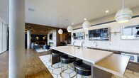 This sophisticated single-floor space seamlessly blends a fully equipped bar and lounge into one open, cohesive environment. The centerpiece is a generously sized white quartz bar island outfitted with multiple draft beer taps and a dedicated sink, surrounded by sleek black-and-gold upholstered bar stools that add a refined, contemporary touch. Three striking white-and-gold spiral pendant lights hang above the island, providing focused illumination and serving as a design focal point.

The bar area is backed by white subway tile and complemented by a wall-mounted flat-screen television, making it well-suited for social gatherings. Recessed lighting and carefully placed sconces contribute to a warm, inviting atmosphere throughout. The flooring transitions between a geometric mosaic tile beneath the bar island and richly patterned parquet wood flooring that extends into the adjoining lounge area.

The lounge zone features a stone-clad fireplace, comfortable seating, and an ornate ceiling detail with ambient track lighting, creating a warm, intimate retreat. Together, the spaces deliver a polished, upscale ambiance ideal for communal entertaining.