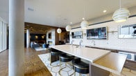 This sophisticated single-floor space seamlessly blends a fully equipped bar and lounge into one open, cohesive environment. The centerpiece is a generously sized white quartz bar island outfitted with multiple draft beer taps and a dedicated sink, surrounded by sleek black-and-gold upholstered bar stools that add a refined, contemporary touch. Three striking white-and-gold spiral pendant lights hang above the island, providing focused illumination and serving as a design focal point.

The bar area is backed by white subway tile and complemented by a wall-mounted flat-screen television, making it well-suited for social gatherings. Recessed lighting and carefully placed sconces contribute to a warm, inviting atmosphere throughout. The flooring transitions between a geometric mosaic tile beneath the bar island and richly patterned parquet wood flooring that extends into the adjoining lounge area.

The lounge zone features a stone-clad fireplace, comfortable seating, and an ornate ceiling detail with ambient track lighting, creating a warm, intimate retreat. Together, the spaces deliver a polished, upscale ambiance ideal for communal entertaining.