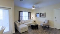 This single-story space features a welcoming living room finished with warm, neutral tones that create a calm and inviting atmosphere. Dark wood-style flooring runs throughout, providing a rich contrast against the light-colored walls. The room centers around a generous white sectional sofa paired with a tufted round leather ottoman, offering both comfort and a classic aesthetic. A ceiling fan with a built-in light fixture is centrally mounted, contributing to both air circulation and ambient lighting. Natural light filters in through a window dressed with layered curtains — a sheer inner panel paired with darker drapes — adding depth and softness to the space. A floor lamp positioned in the corner supplements the natural light during evening hours. Wall décor includes framed equestrian-themed artwork, lending a refined, traditional character to the room. A patterned accent chair and small side table round out the seating area, adding visual interest and functionality. Overall, the space combines practical design with tasteful, cohesive décor for a comfortable living environment.