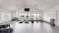 Located at 2500 Central Park Ln in College Station, Texas, this fitness center is a thoughtfully designed exercise facility housed within a single-floor space. The room features a striking vaulted ceiling that lends an open, airy feel, complemented by recessed lighting and large, multi-pane windows along the front wall that flood the space with natural light and offer views of the surrounding outdoor area, including what appears to be a pool and neighboring structures.

The layout is divided into distinct functional zones. A dedicated turf area with bright green artificial grass provides space for functional training, accommodating battle ropes, a tire flip sled, medicine balls, resistance bands, and a weight bench. Adjacent to the turf, a full rack of dumbbells offers a wide range of free weight options alongside a cable/multi-functional strength machine for resistance training.

The cardio section features multiple treadmills, stationary bikes, and an elliptical trainer arranged to face the expansive front windows. A wall-mounted flat-screen television is positioned above the window bank for entertainment during workouts. The flooring throughout the non-turf areas consists of durable, dark rubberized material suited for high-traffic fitness use. The clean, white walls and bright interior create a motivating and modern training environment.